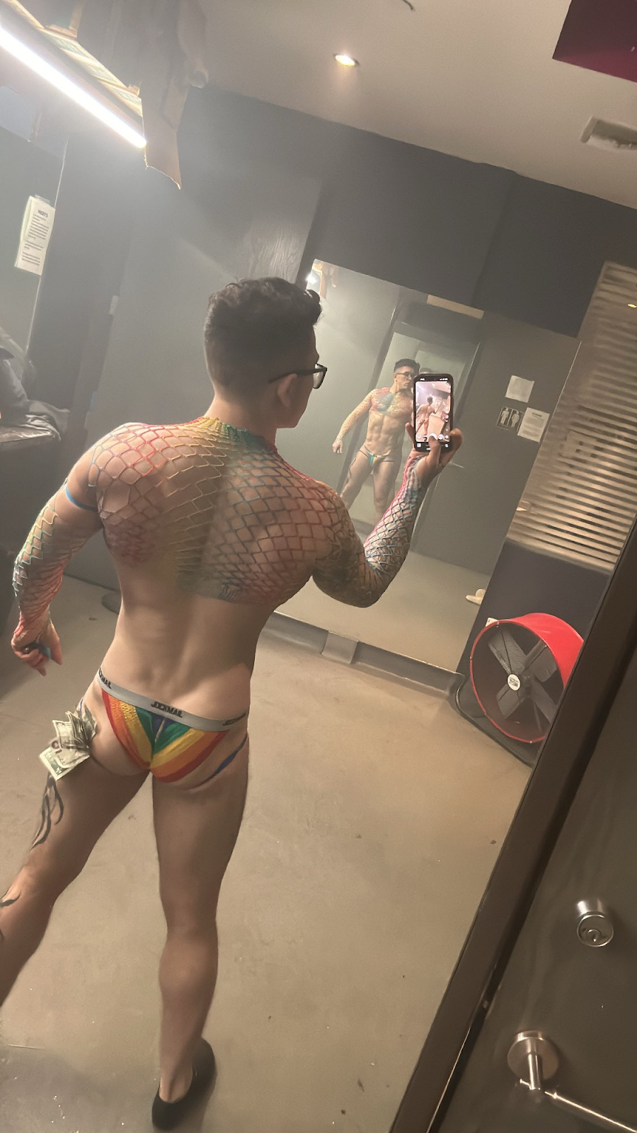 stripper and onlyfans content creator Clark Davis wearing rainbow jockstrap and dollar bills in his underwear wearing mesh shirt and taking selfie in stripclub mirror