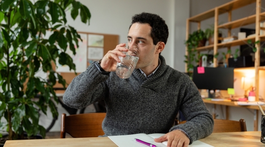Homme assis à son bureau portant un gilet gris et buvant un verre d'eau