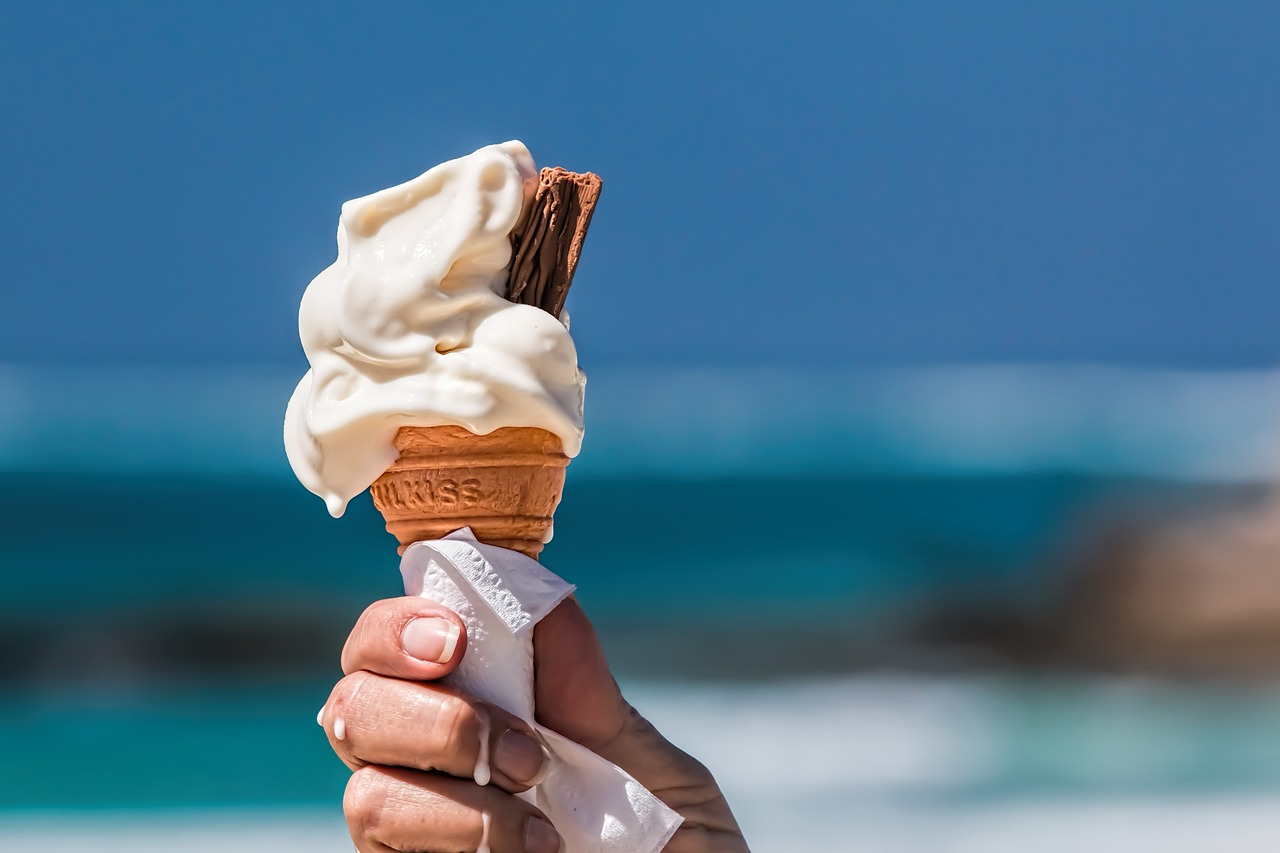 A hand holds a vanilla ice cream cone at the beach.
