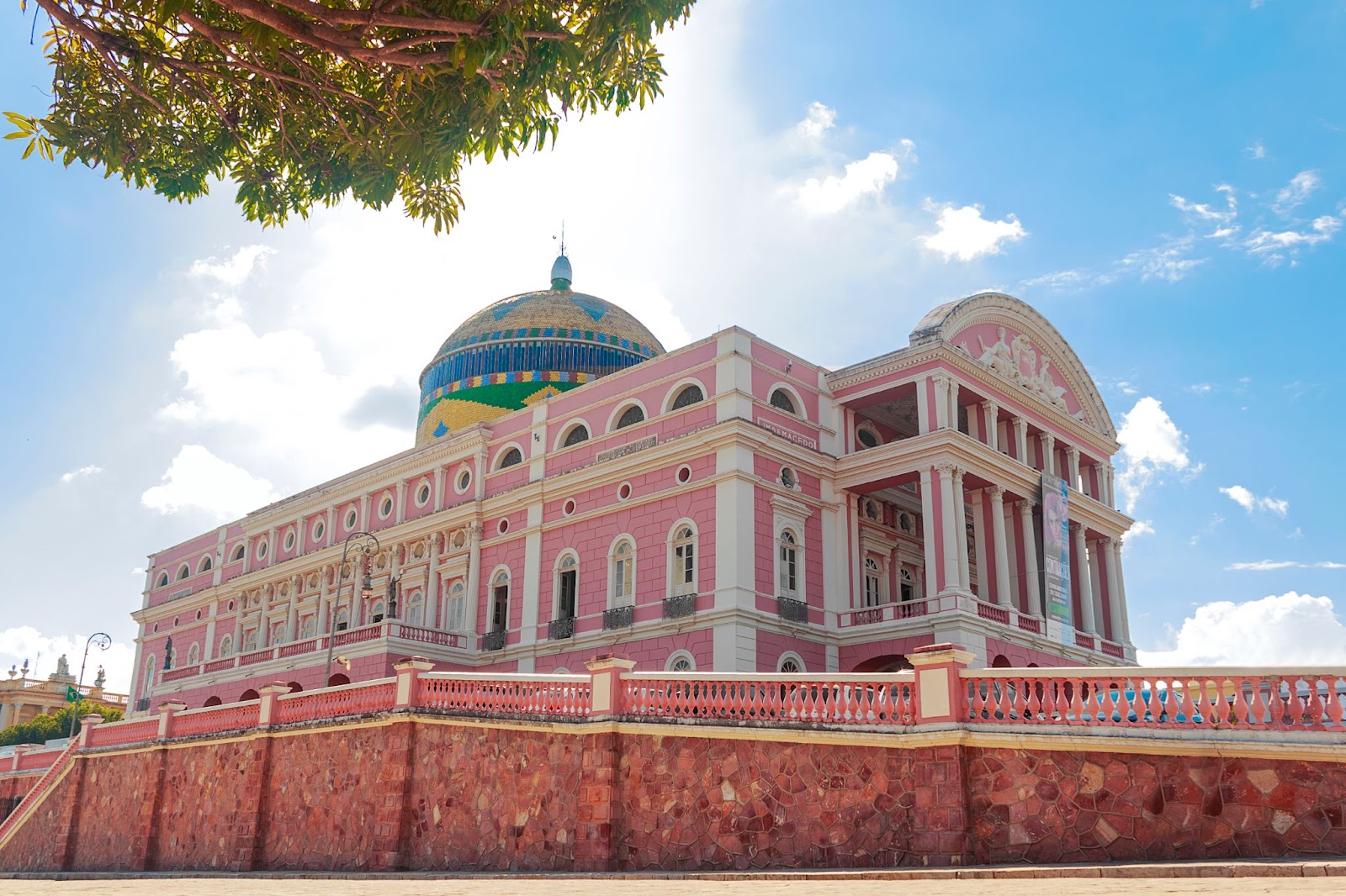 Teatro Amazonas, em Manaus. A grande construção de arquitetura renascentista tem uma fachada de cor rosa marcada por grandes colunas e muitas janelas. No topo, há uma cúpula com mosaicos em azul, verde e amarelo