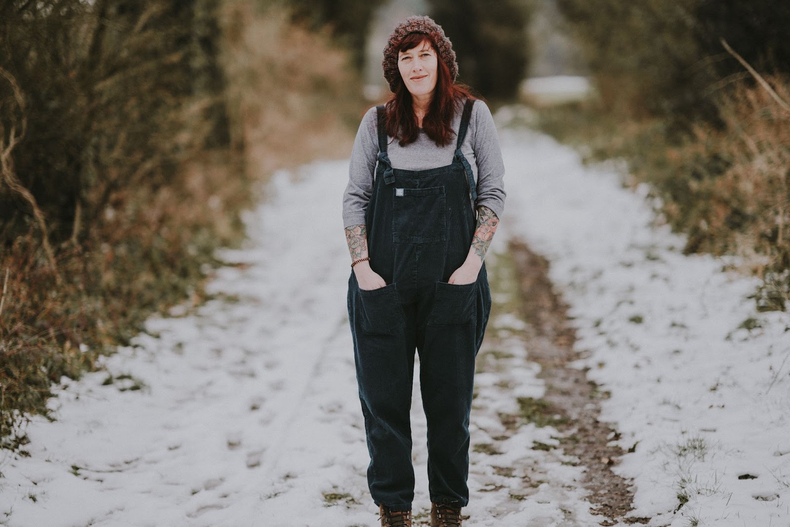 a woman standing on a dirt road with snow on it, wearing a knitted hat and coveralls, and a shirt with her forearms exposed to show two tattoo sleeves