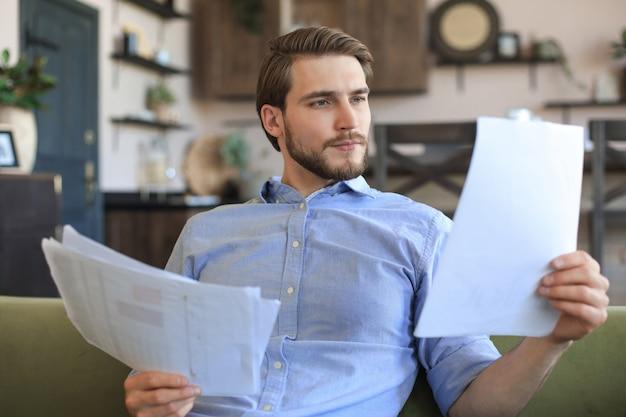 Concentrated young freelancer businessman sitting on sofa with laptop and examining documents.