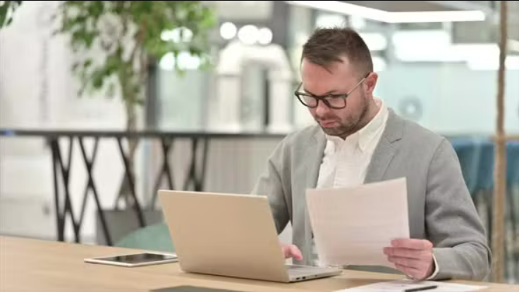 En mand sidder bag papirer og bærbare computere på kontoret