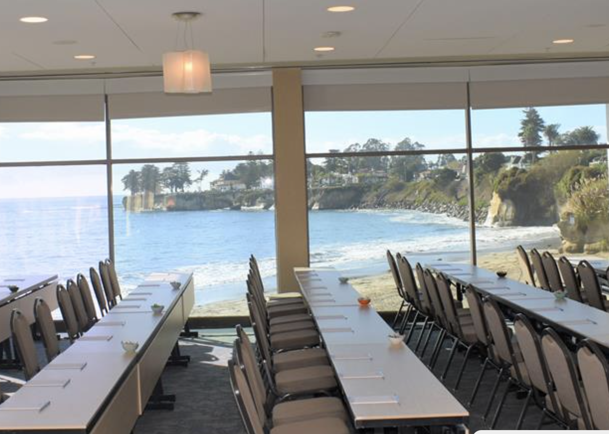 Photograph of Dream Inn conference room. Rows of long tables with chairs, next to large window overlooking the ocean and beach.