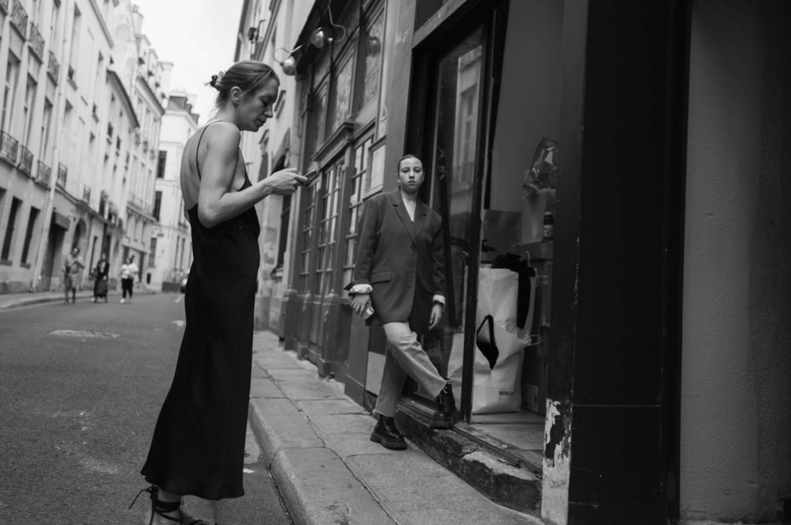 Black and white photograph of two women standing in the streets of France, one wearing a black slip dress looking at her phone, the other standing off to the side with a dark blazer coat and slacks.