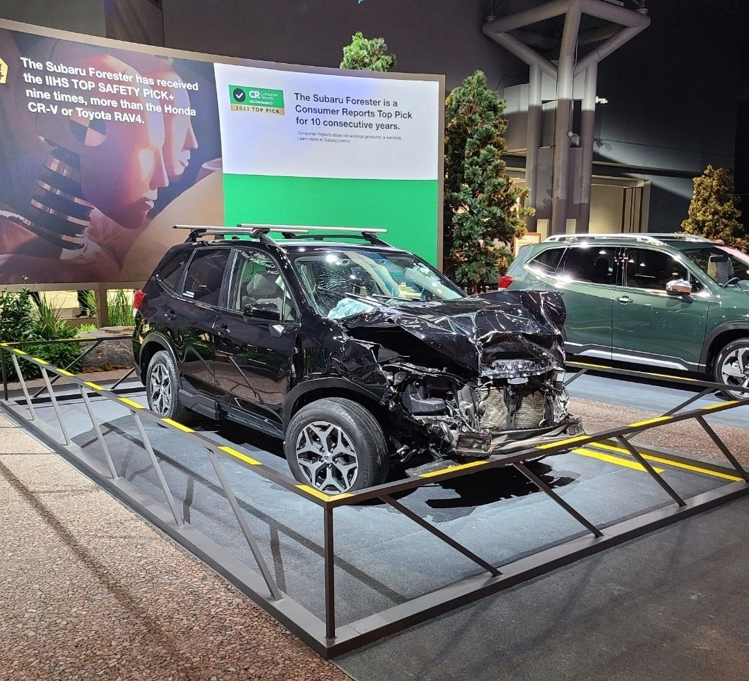 A picture of Subaru's booth at the NY Auto show. The highlight is a Subaru that's been in a seemingly very bad accident. The front is completely crumpled in, but the seating area, where the people would be, is more or less okay. The copy on the board behind it says "The Subaru Forester is a Consumer Reports Top Pick for 10 consecutive years."