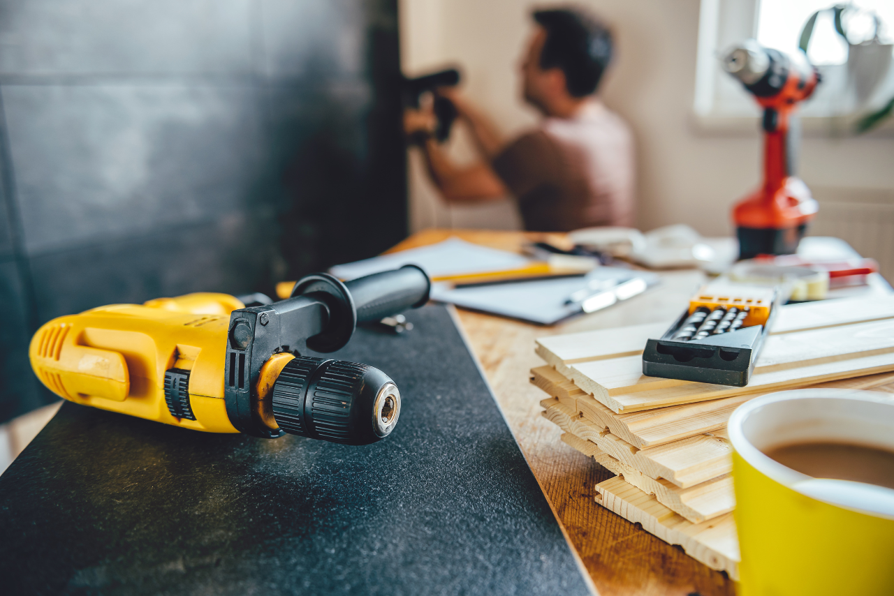A power tool sits in the foreground of the image while an HVAC technician works in the background.