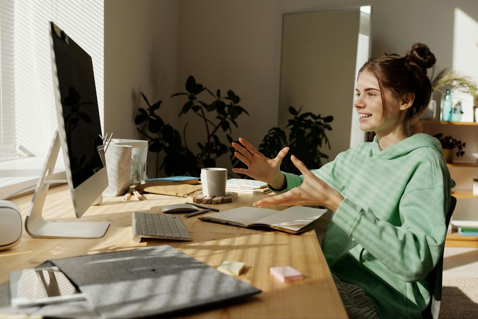 A lady in a mint green hoodie and space bun-styled hair, sitting and smiling at her computer and making gestures with her hands as if talking to her audience online.