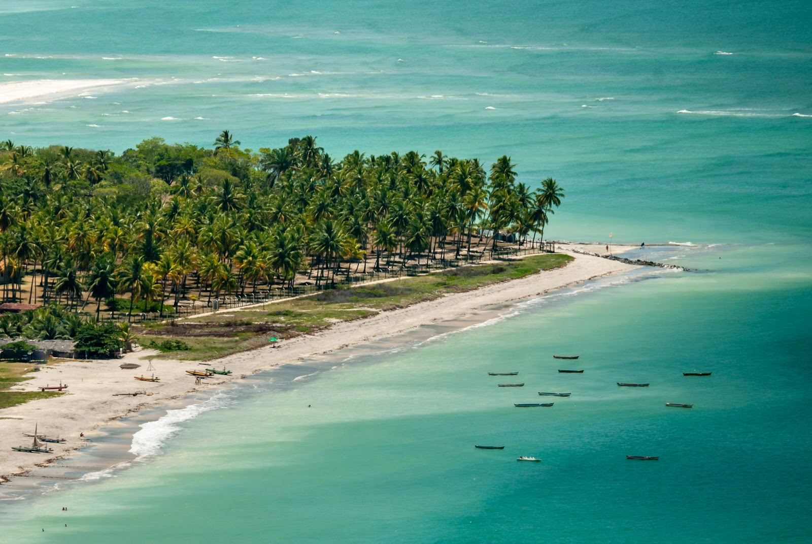 Foto aérea de parte da Ilha de Itamaracá. Pontal banhado pelo mar azul claro, com faixa de areia branca, gramado e muitos coqueiros verdes