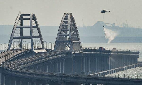 A view of a large bridge, part of which is smoldering as a helicopter drops water onto that section.