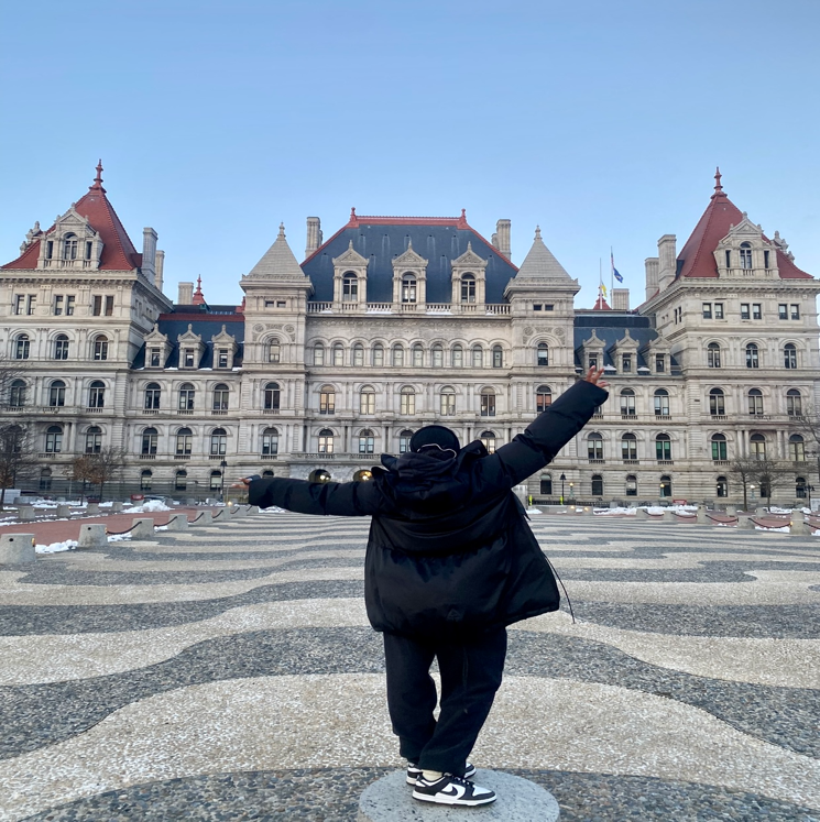 A person standing in front of a large building with New York State Capitol in the background  Description automatically generated