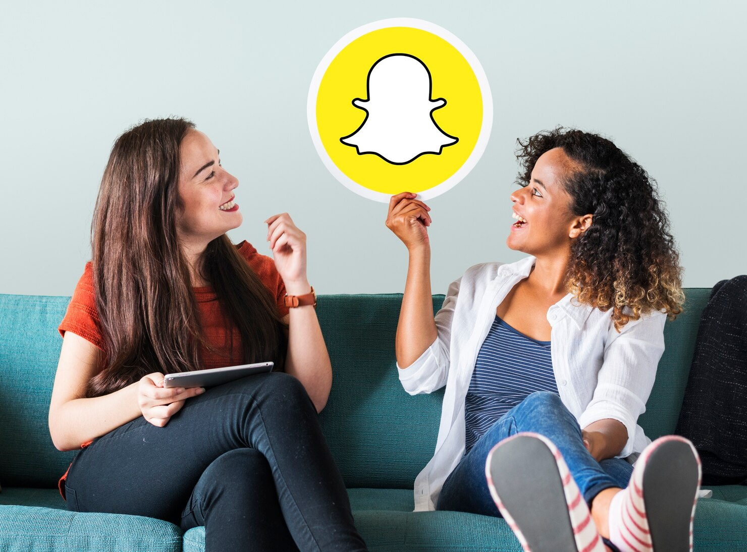 Two women laughing while discussing good captions for snapchat and holding a Snapchat icon.
