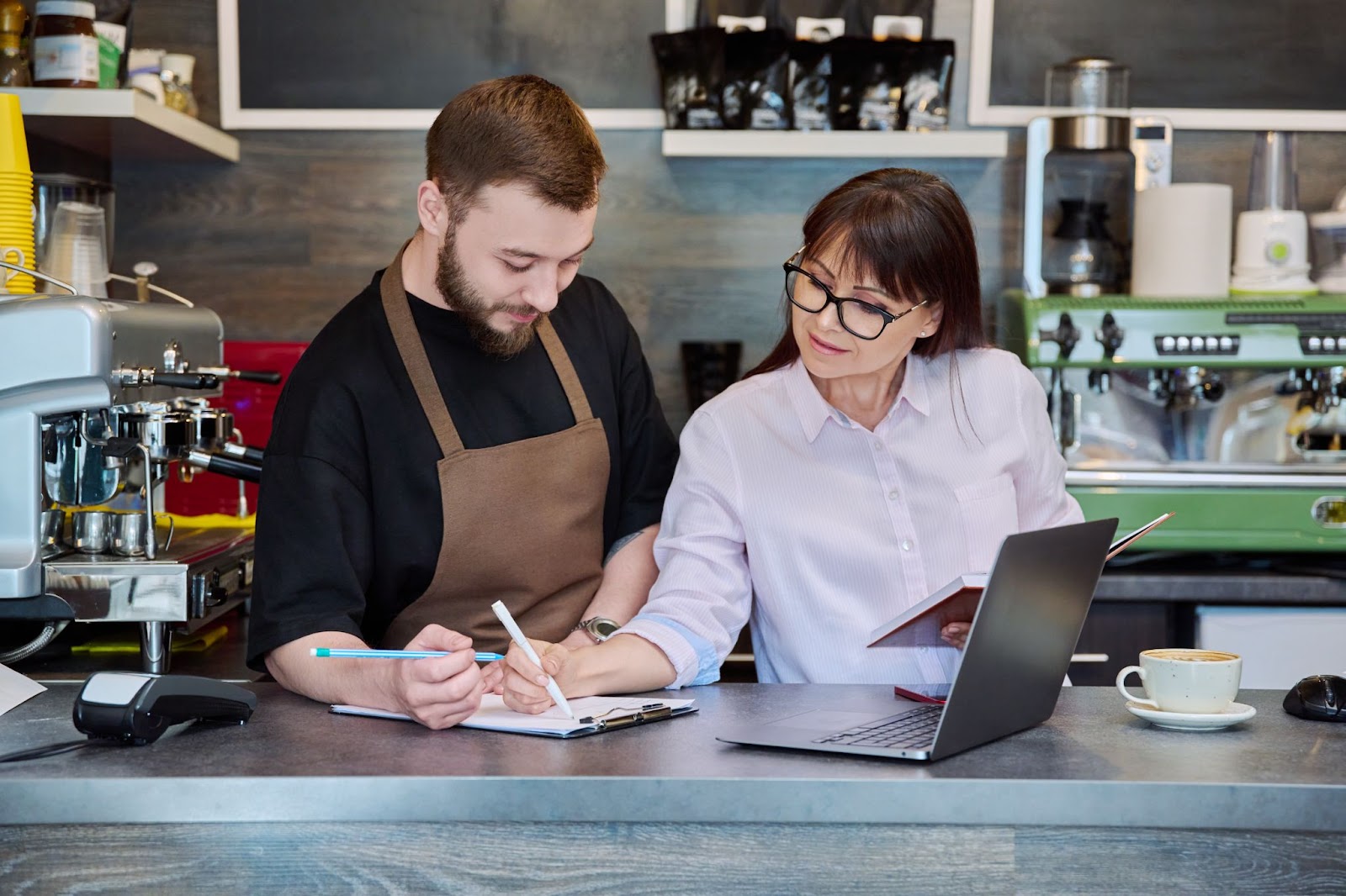 A man and a woman collaboratively working on a laptop at a coffee shop table, with coffee machines and neatly arranged cups on shelves in the background.