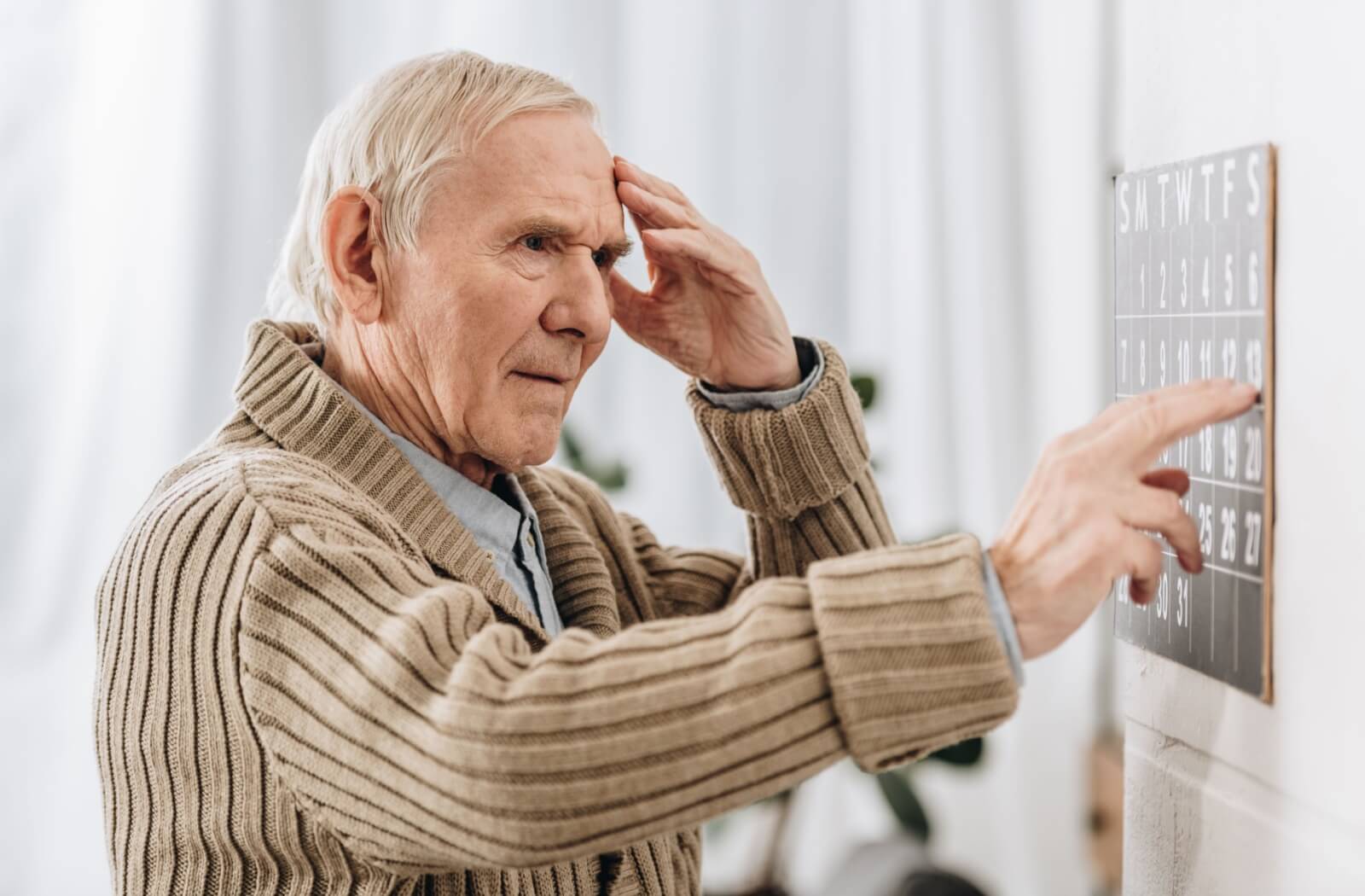 An older adult man in his sweater trying to remember something as he points to a day on a calendar.