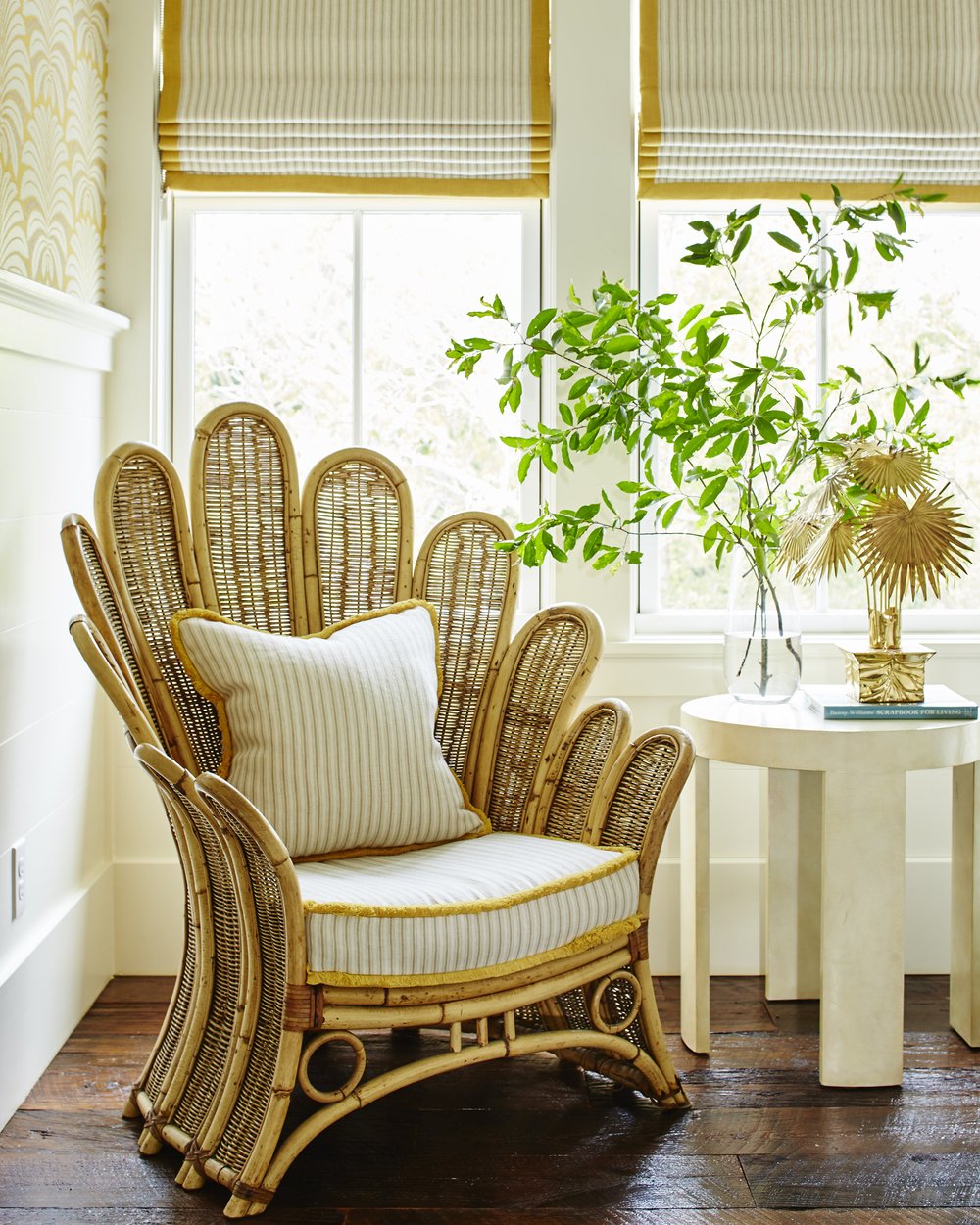 A bright, natural-light-filled reading corner with a flower rattan chair, roman shades upon the windows, and a round side table adorned with plants and other decoration.