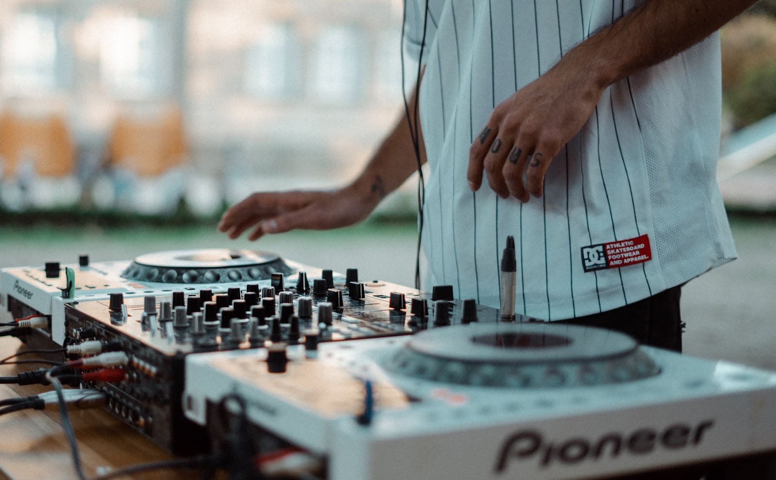 A close-up of a DJ's hands adjusting knobs on a Pioneer DJ mixer at an outdoor event, set against a casual, daylit backdrop.