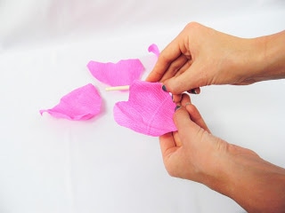Pink petal pieces lay on a white table in the background, while in the foreground, Abbi Kirsten's hands show how to curl the petals already attached to the stem with a thin dowel. Crepe paper flowers look more realistic and are fun to make. 