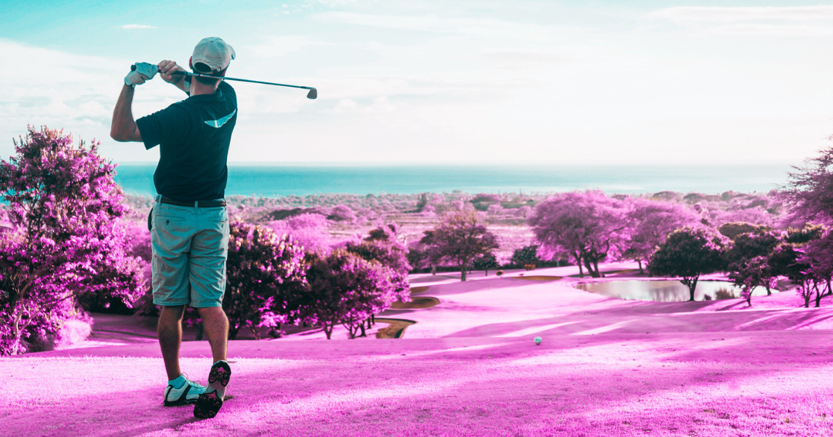 A golfer wearing a blue shirt and white cap is captured at the end of their swing, highlighting both form and motion. The red-toned ground contrasts well with their attire to emphasize action.