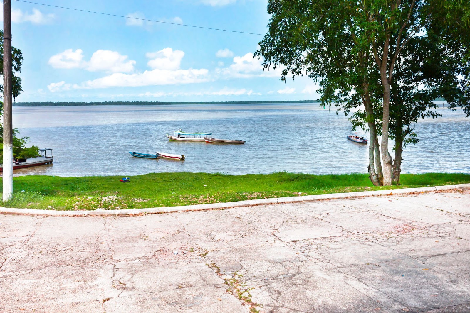 Parte da Orla de Icoaraci. Um estreito gramado verde separa a calçada da orla e a água do rio, que reflete o azul do céu. Algumas embarcações de pequeno porte flutuam pelas águas