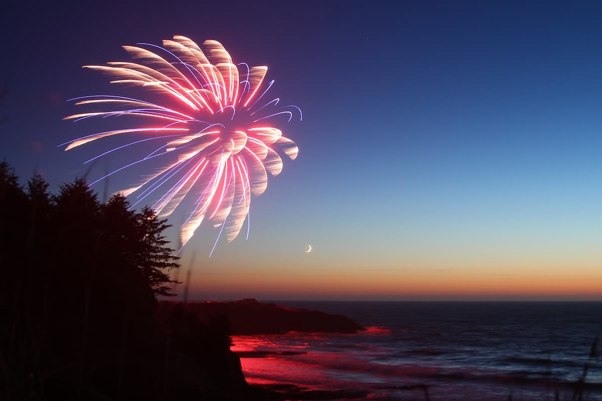 Purple and orange firework over Pacific Ocean along the Oregon coast with moon in background