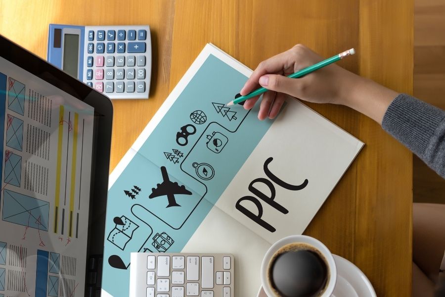 Person seen from above, drawing marketing symbols on a "PPC" (Pay-Per-Click) labeled paper next to a laptop displaying charts.