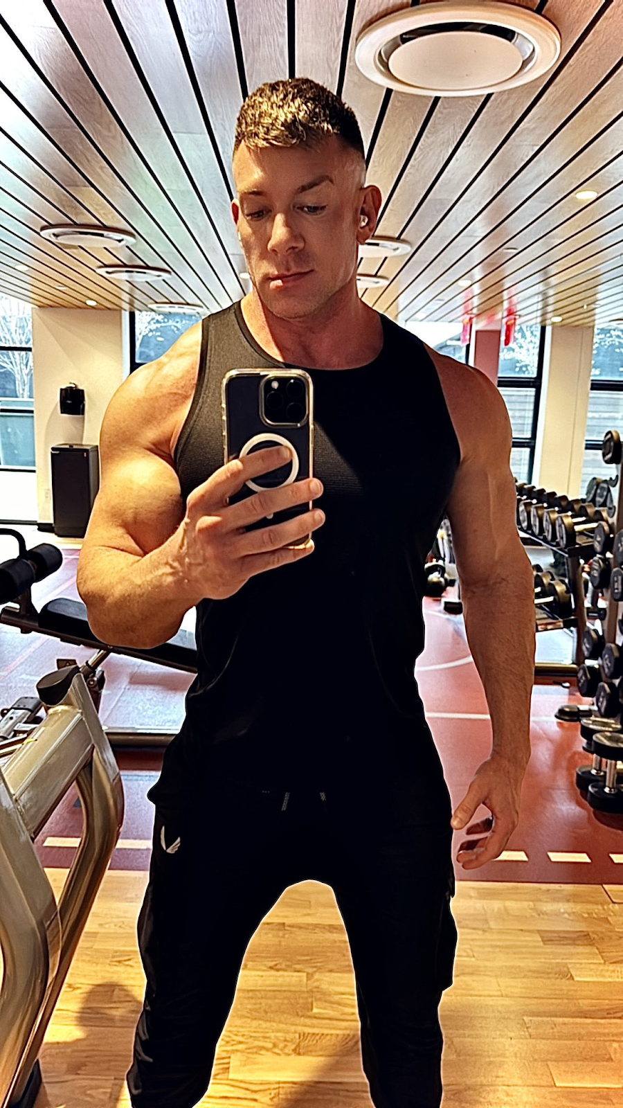  Bruce Beckham taking an iphone mirror selfie in black tank top showing off bulging biceps in front of gym weight rack