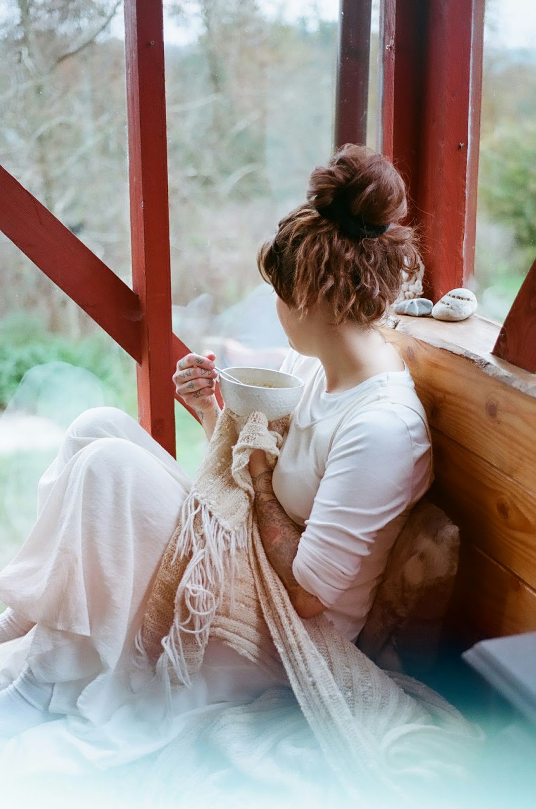 Frau sitzt mit Teetasse und Decken in geschlossener Terrasse und beobachtet Garten. 