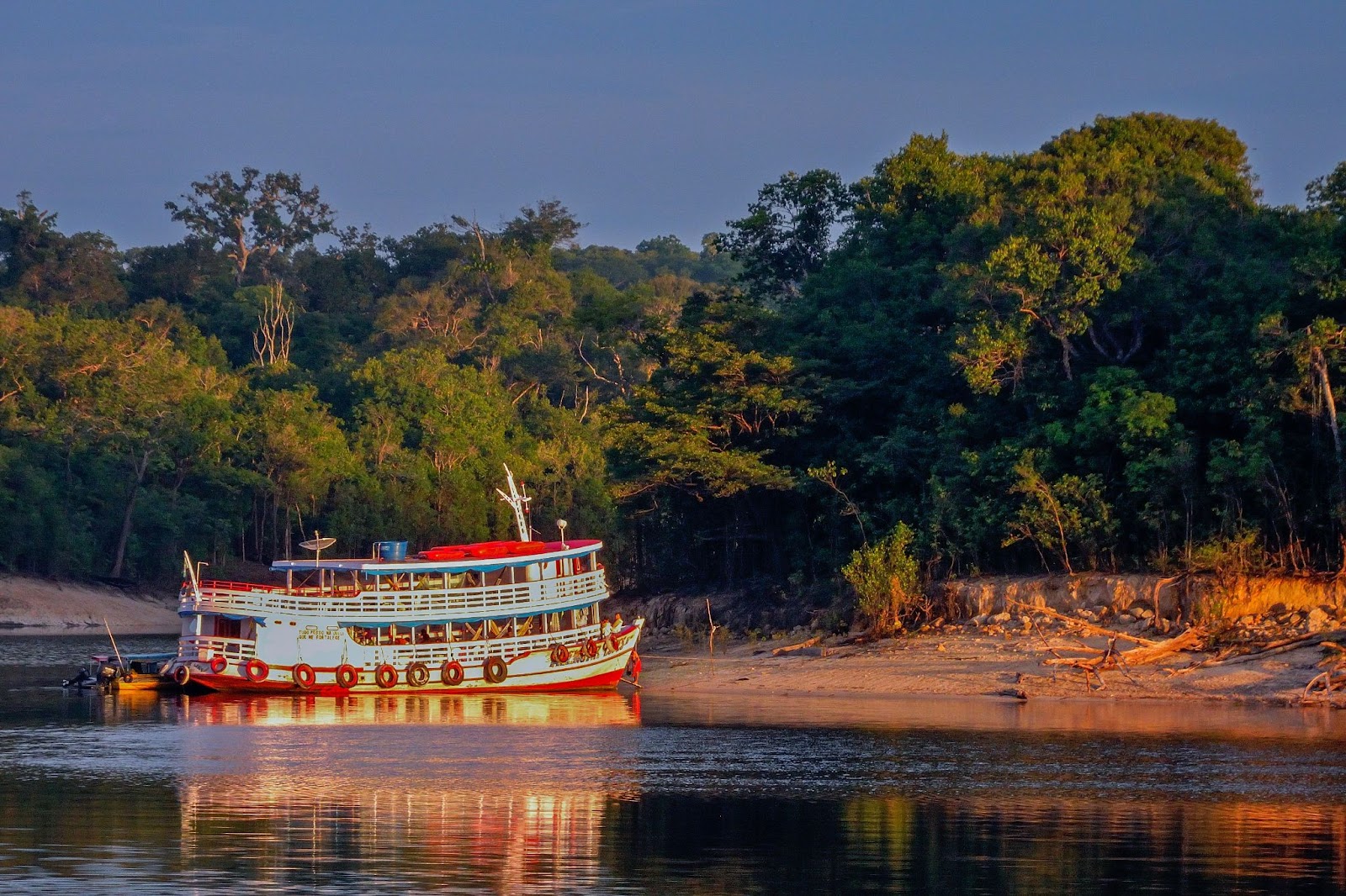 Embarcação de dois andares flutuando no rio em Manaus. Ela se aproxima da terra, onde há um trecho de mata verde e densa