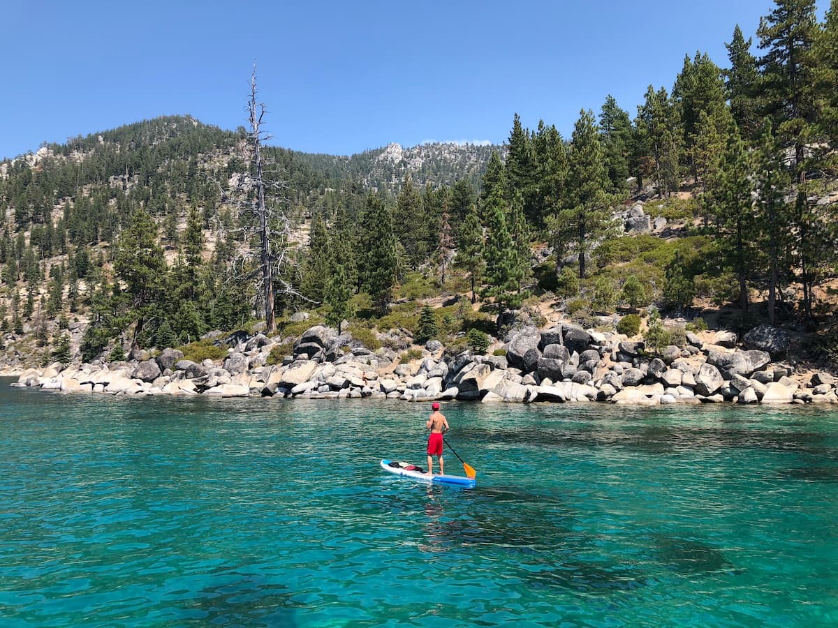 Man stand-up paddle boarding on clear blue waters on lake Tahoe with rocks and trees in background
