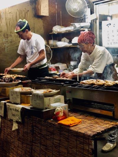 Two men cooking yakitori skewers at a street-side stand.