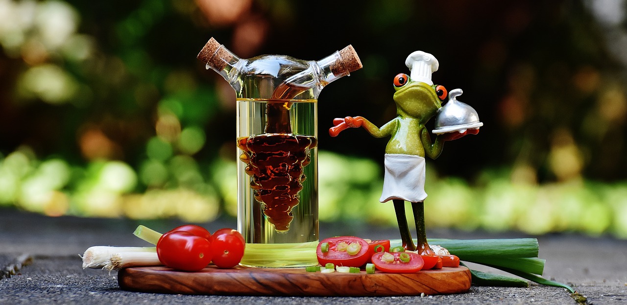A miniature frog dressed as a chef standing over slices of cherry tomatoes and a bottle of apple cider vinegar.