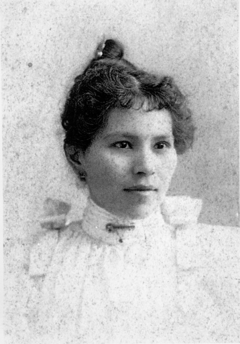 Rachael Blythe Bauer looking to the right of the camera.   She has dark eyes and dark hair.  Her hair has short curly bangs in front and is up in a bun.  She is wearing a high-necked, white shirt with ruffles.