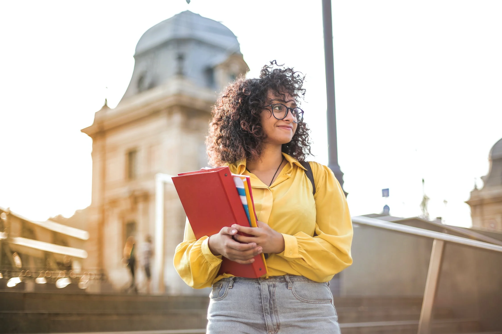 Girl with glasses and a yellow shirt holding books, smiling in front of a building - 10 Things to Complete in Your First Term of College