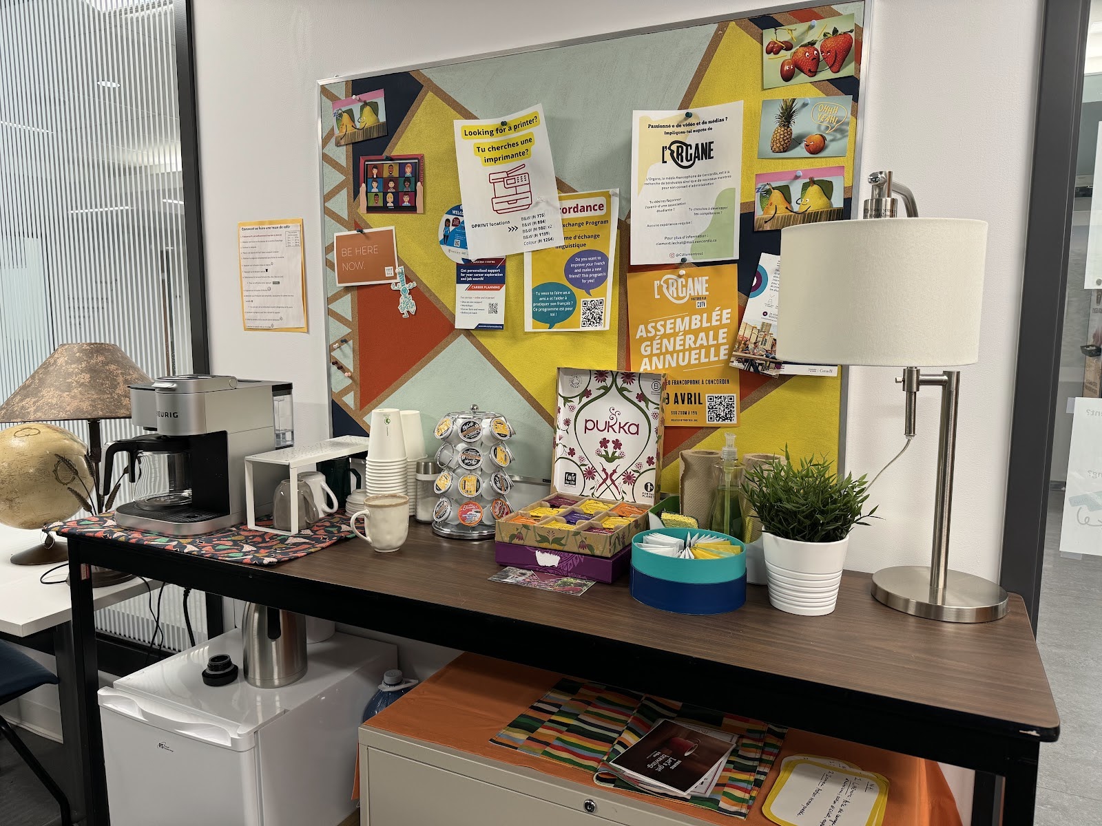 A table with a coffee maker, mugs, coffee pods, and tea sits in front of a colourful corkboard with various flyers and posters pinned to it.