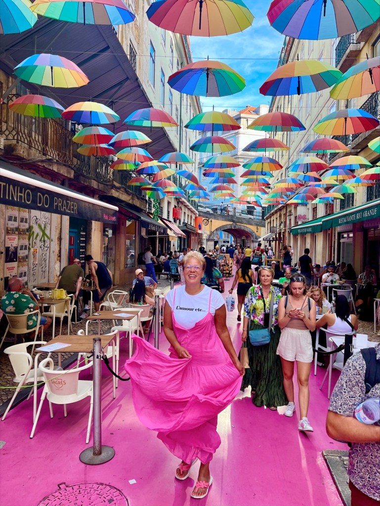 Rachel Boesing smiles brightly as she stands outside near tables from various restaurants where people are eating. Above her are lots of tiny, colorful umbrellas. She's moving, making her pink dress "whoosh"
