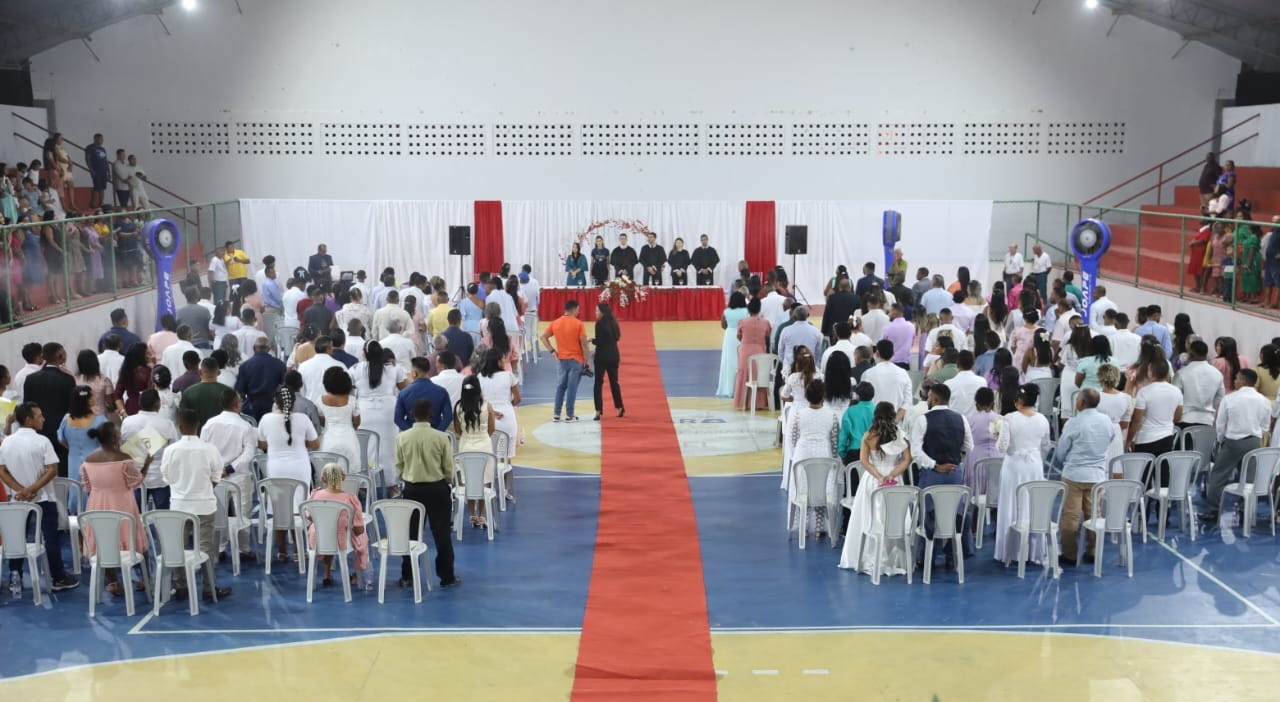 FOTO HORIZONTAL, COLORIDA, DE CASAIS DE PÉ DIANTE DE CADEIRAS BRANCAS, EM AUDITÓRIO DURANTE CERIMÔNIA DE CASAMENTO COMUNITÁRIO. ENTRE AS FILEIRAS DE CADEIRAS, TAPETE VERMELHO. AO FUNDO, QUATRO PESSOAS DE PE, VESTIDAS DE TOGA RPETA.