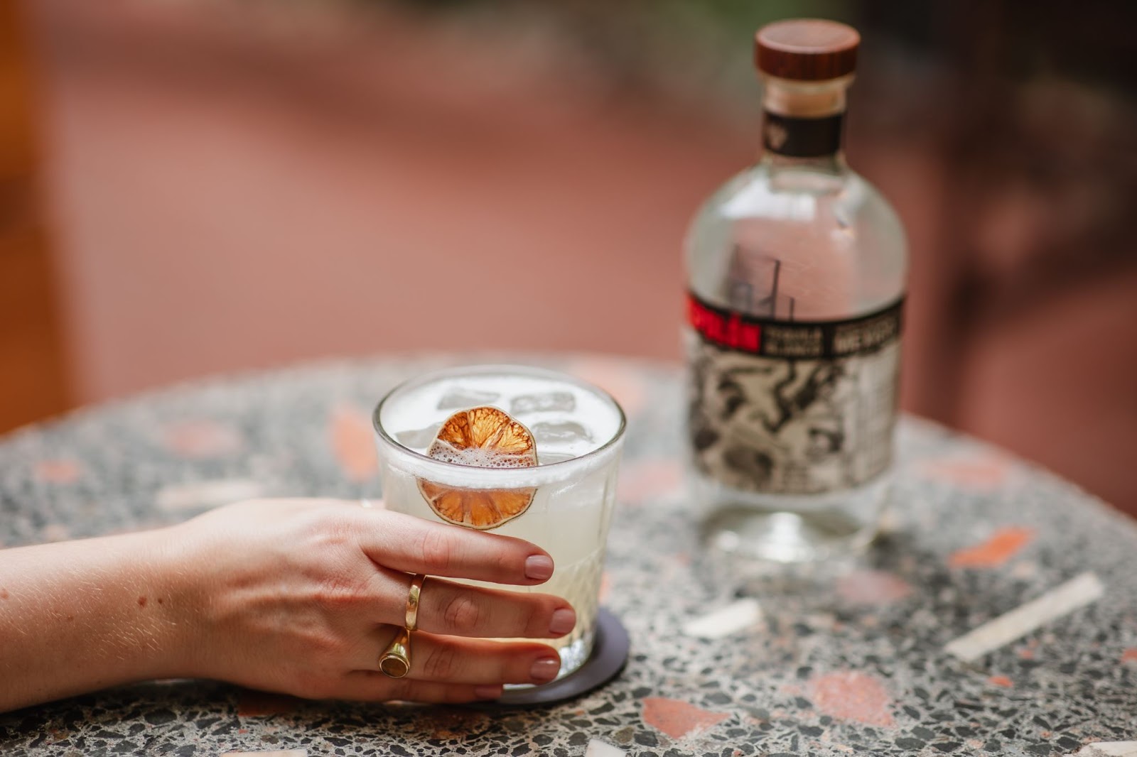 A hand holds a glass with margarita in it, next to a bottle. 