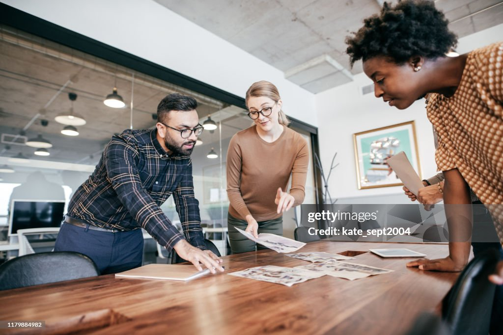 Three colleagues collaborating over documents and photographs spread out on a table in a office space.