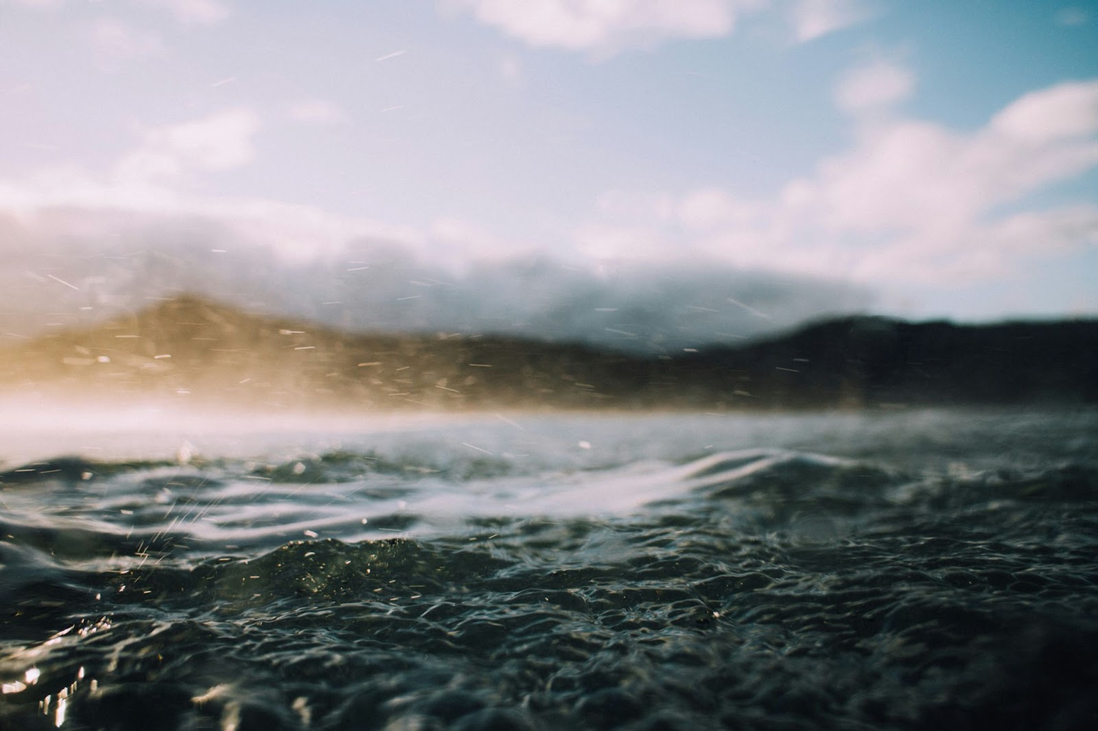 Close up picture of a lake with choppy waves and spray being kicked up by wind and out of focus mountains in the background.