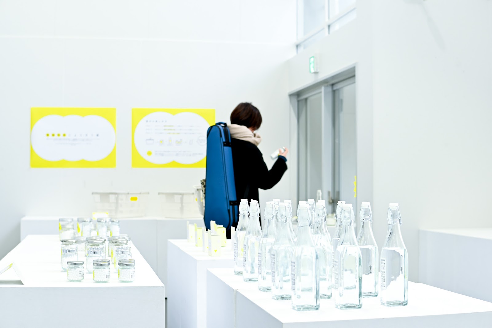 婚姻制度をテーマにした展示会会場の写真。白い空間に、黄色いデザインのパネルが貼られ、展示台には様々な形の瓶が並べられている。