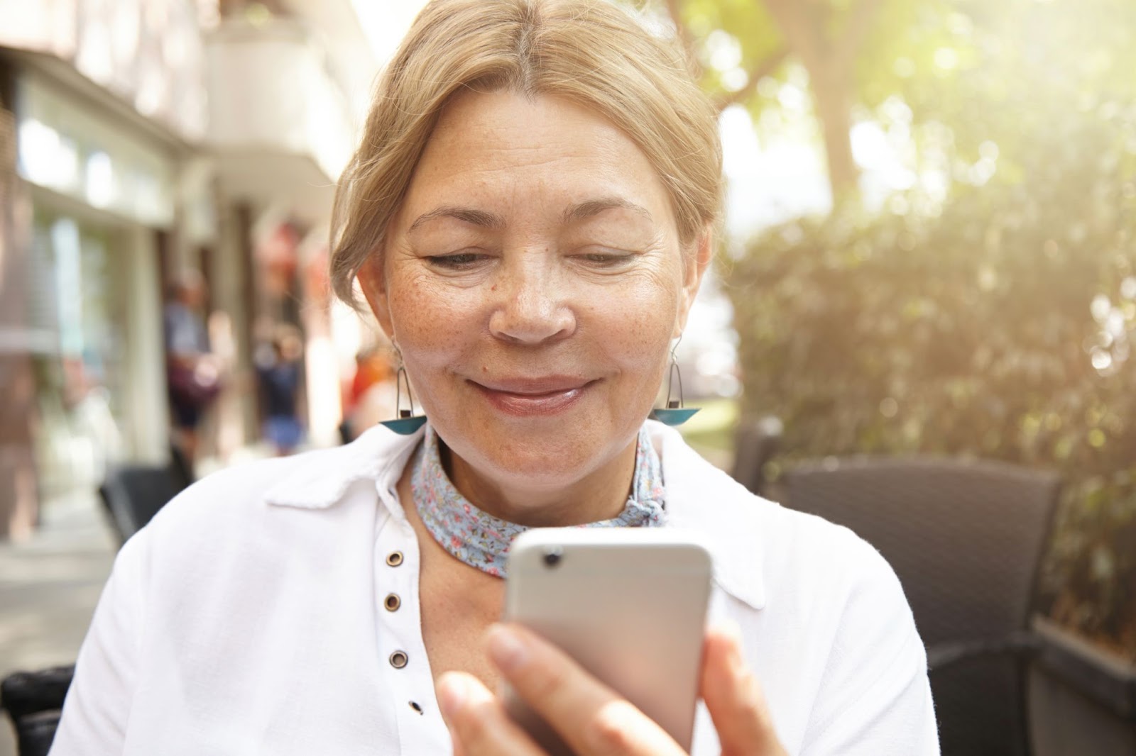 Imagem de uma mulher de meia-idade observando a tela de seu celular. Ela sorri, usa cabelos presos, um lenço em seu pescoço e veste blusa branca. 