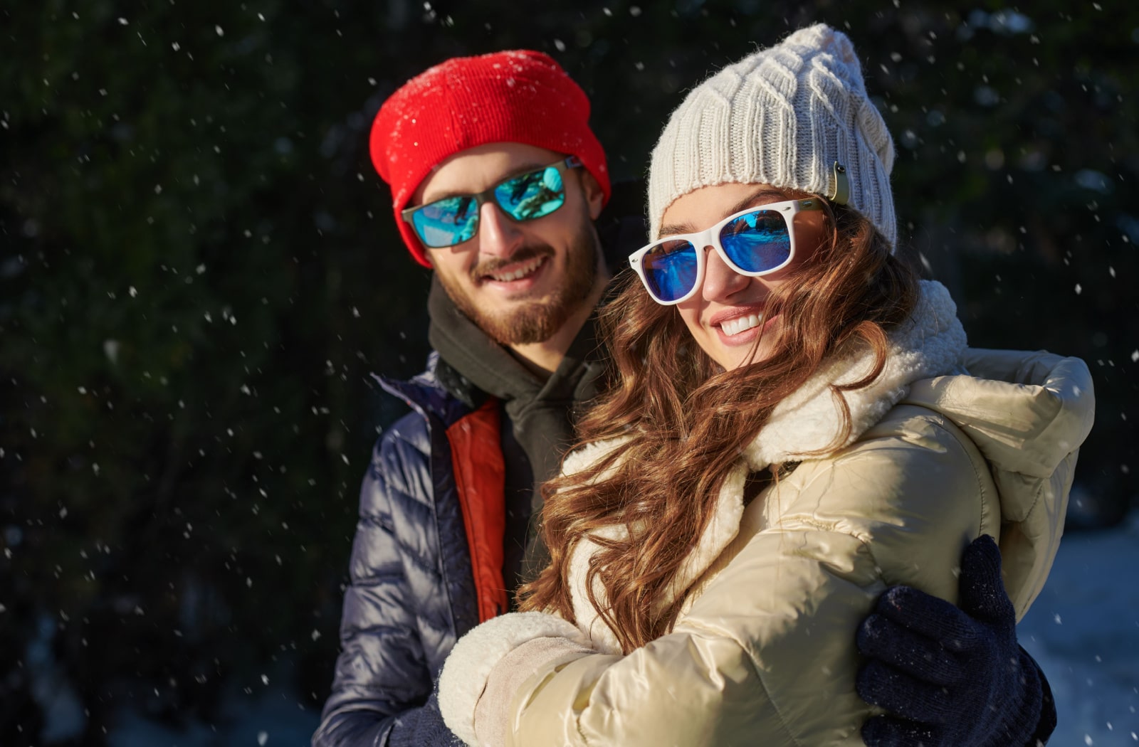 A couple standing together outside in the snow, wearing winter jackets, hats, and sunglasses