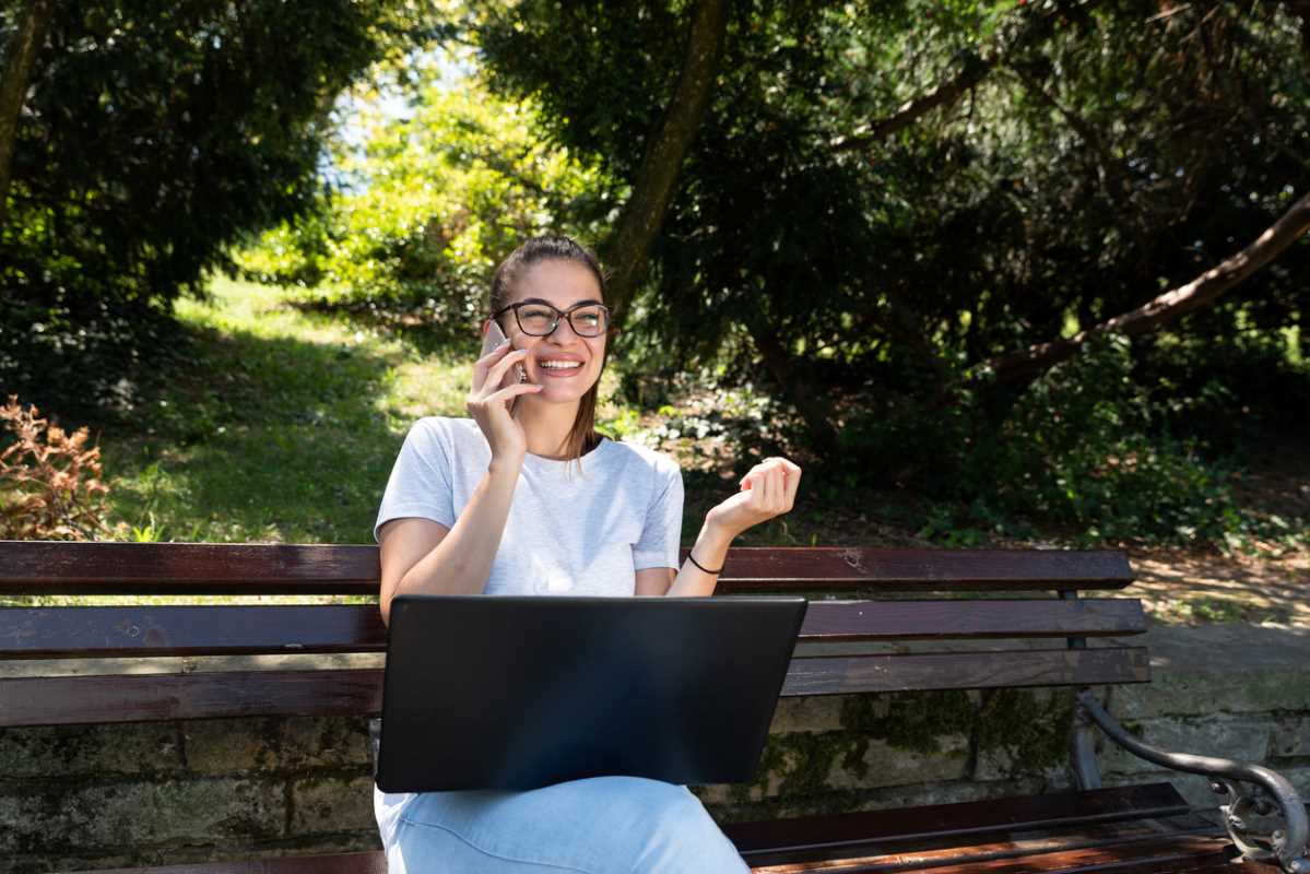 A young freelance wedding planner with reading glasses and a laptop sits on a park bench.