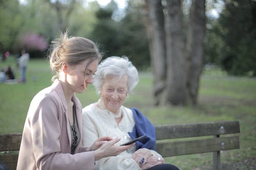 Free Side view of smiling adult female helping aged mom in using of mobile phone while sitting together in park Stock Photo