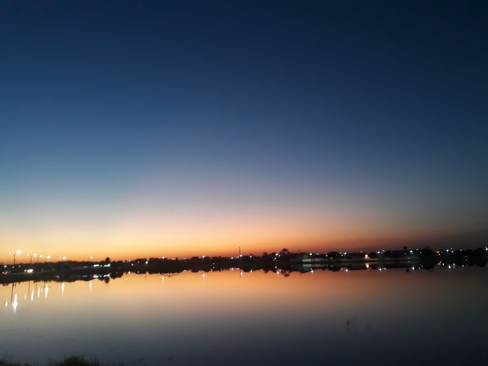 Pôr do sol sobre o Açude Grande, em Campo Maior. Por causa da chegada da noite, o céu está dividido entre tons de azul e laranja. A água do açude reflete os pontos luminosos da iluminação noturna da cidade.