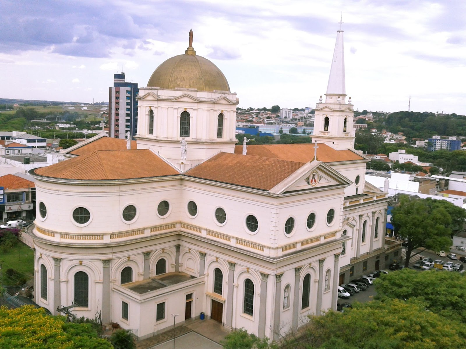 Imagem aérea da parte traseira da Basílica Santuário Santo Antônio de Pádua. Igreja com árvores ao seu redor. Ao fundo, é possível ver um pedaço das casas da cidade.