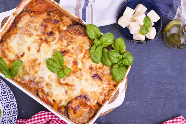 An eggplant lasagna on a baking dish, served on a purple table with cheese cubes arranged on the side