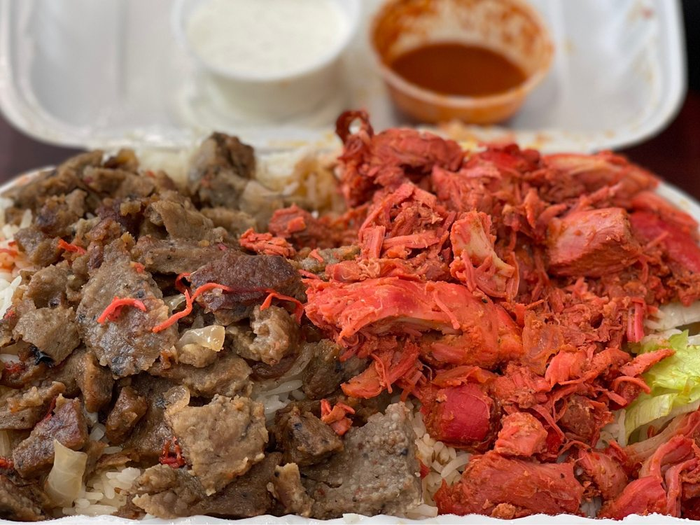 Photo from Mediterranean Halal Food Cart's Yelp page. A close-up of food in a to-go container. On the left is a brown meat that looks like gyros, and on the right is a shredded meat that is bright red in color. Underneath are glimpses of rice and lettuce, and in the back are two open sauce containers.