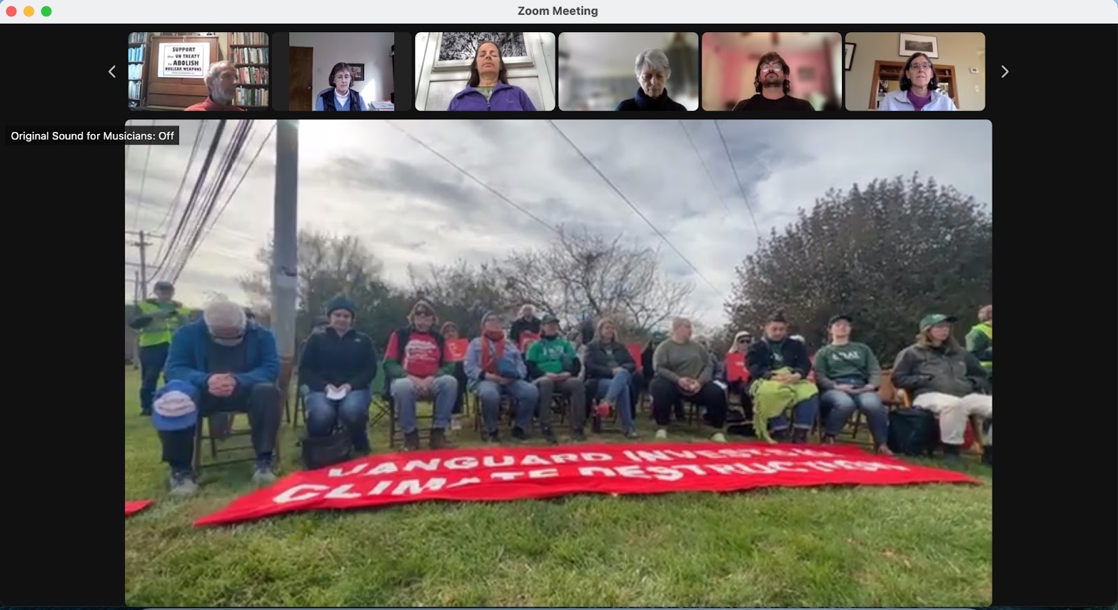 Screenshot of a Zoom video meeting. The largest video is of a group of people seated outside in poses of reflection and with a large red and white protest banner in front of them. Above that large video is a row of smaller videos showing individuals inside, seemingly in their homes, also in poses of reflection. 