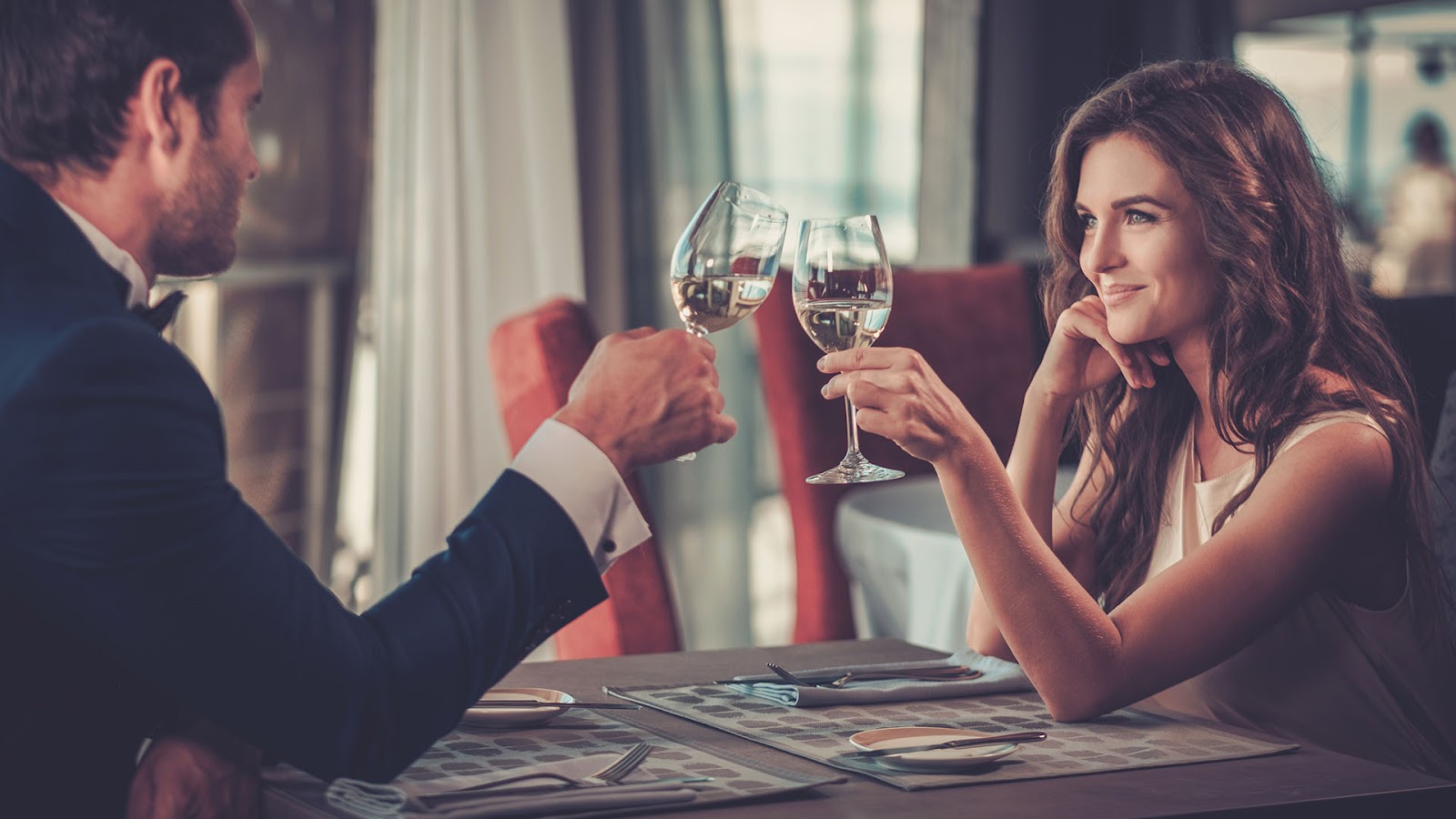 brunette man in a suit toasting his wine glass with a woman sitting across from him